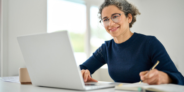 Smiling woman wearing glasses works on a laptop while writing notes at a desk in a bright home office