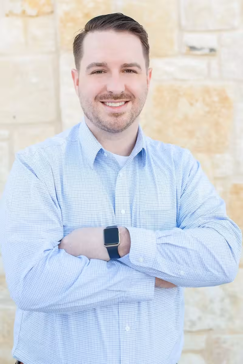 Smiling man with short hair wearing a light blue checkered shirt and a smartwatch, standing with arms crossed in front of a light stone wall.