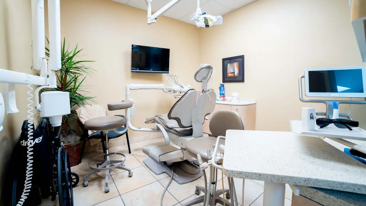 Modern dental examination room with beige dental chair, stools, equipment, a TV on the wall, and a plant in the corner.