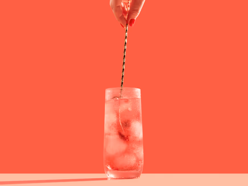 Hand holding a spoon with brown powder moving to pour it into a glass of sparkling water with ice cubes against an orange background.