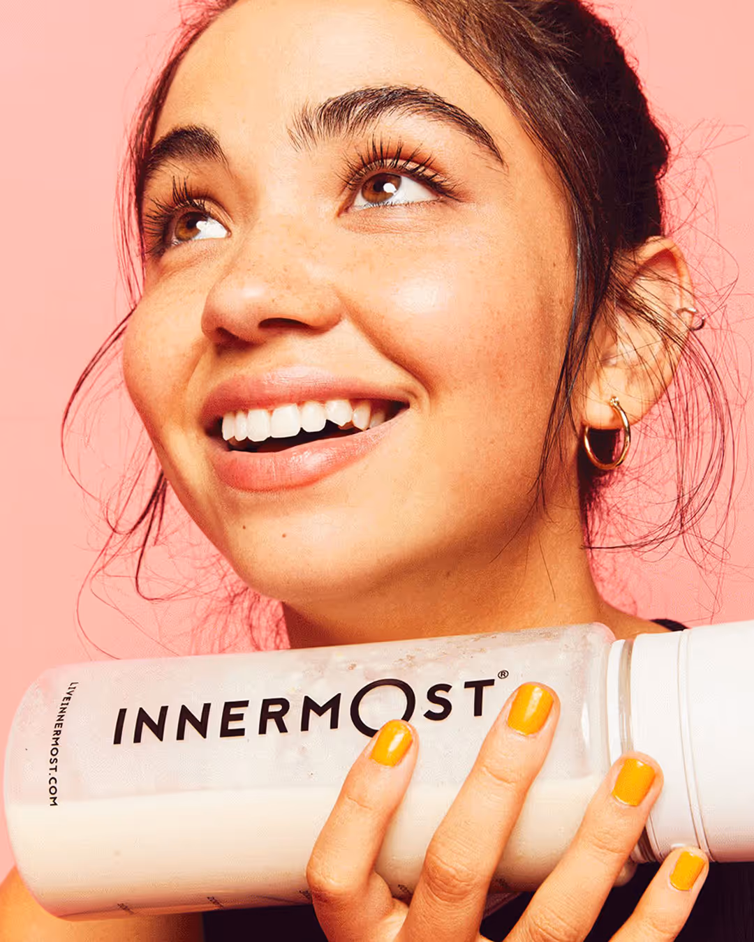 Smiling woman with yellow nail polish holding a white Innermost bottle against a pink background.