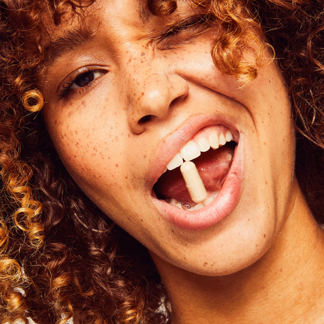 Young person with curly hair and freckles holding a beige capsule pill between their teeth.