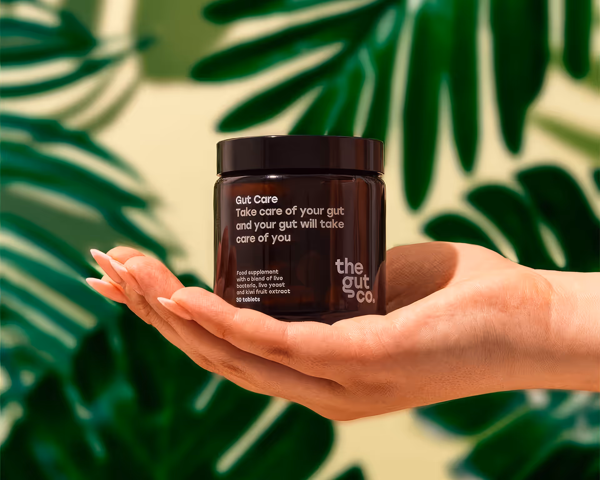 Hand holding a dark jar labeled Gut Care supplement by the gut co against a background with green leaves.