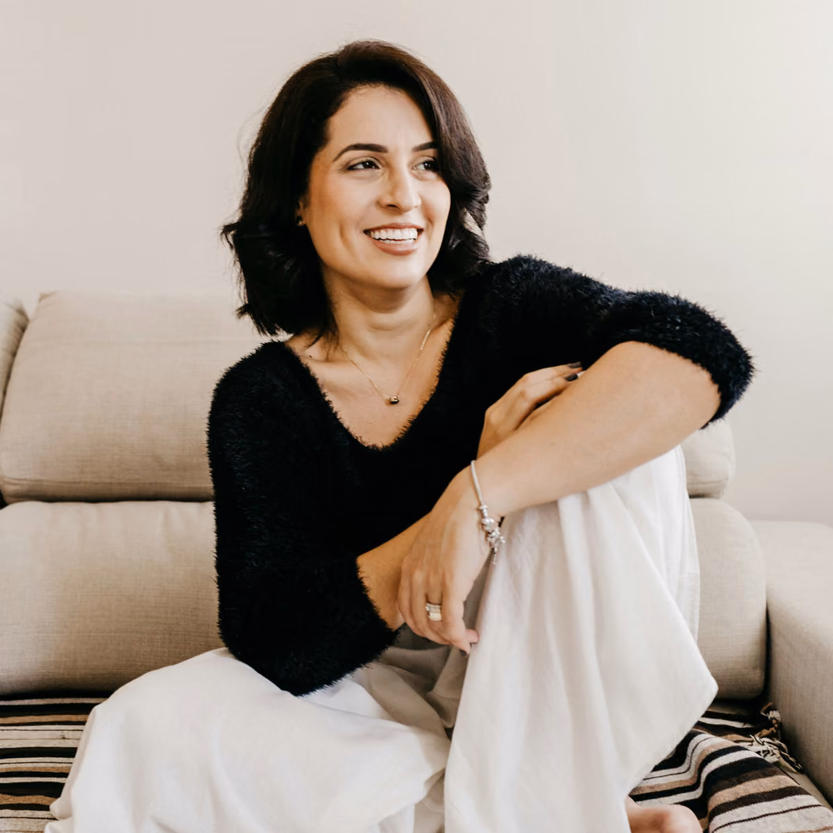 Smiling woman with short dark hair in black sweater and white pants sitting on a beige couch.