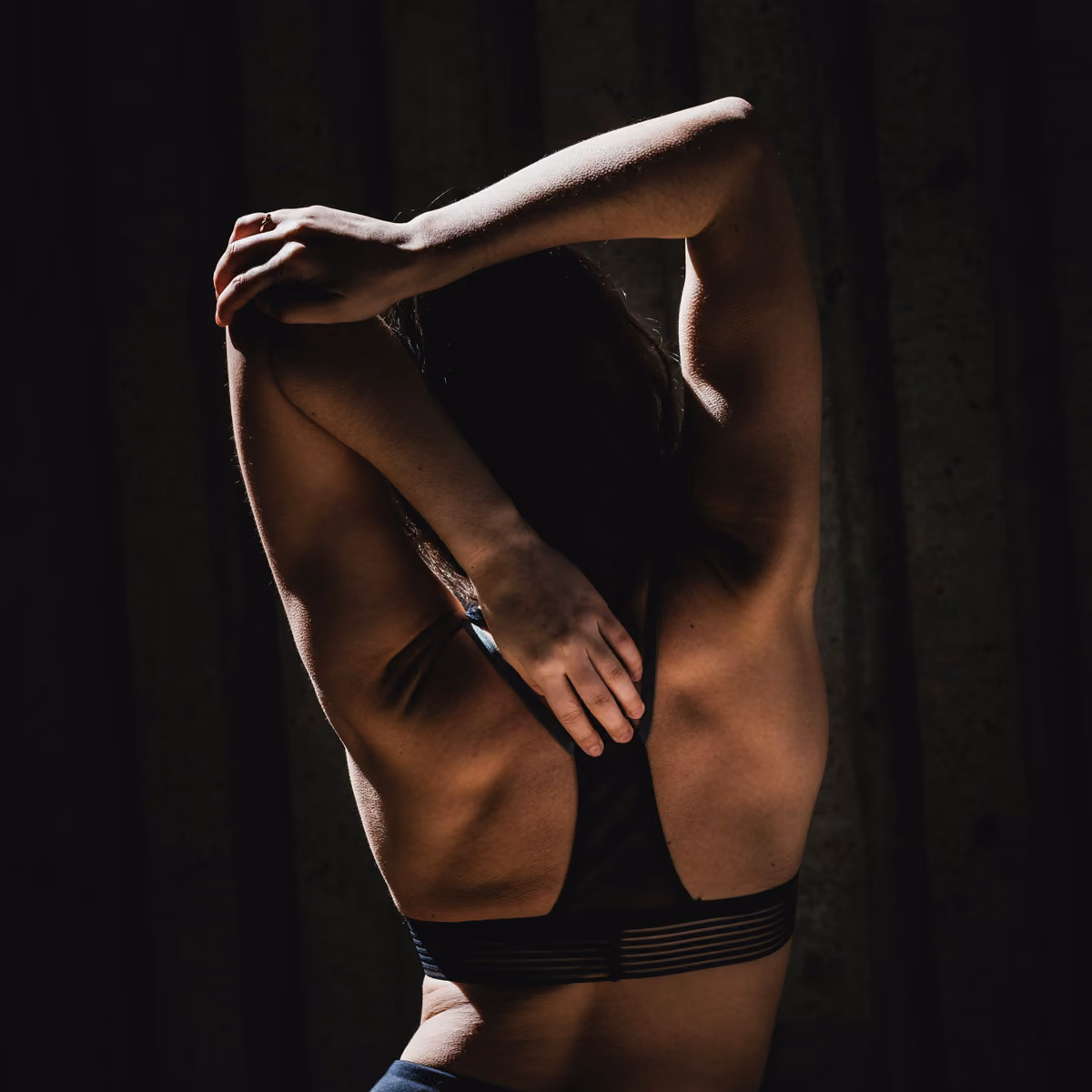 Person in sports bra stretching arms behind head showing upper back and muscles.