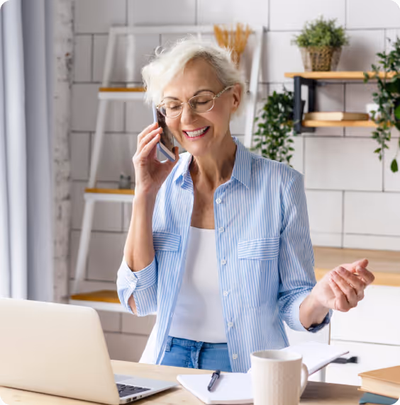 An older lady smiles during a phone call with her energy company, delighted to learn that her monthly bills have decreased significantly, all thanks to the green home improvements she's made.