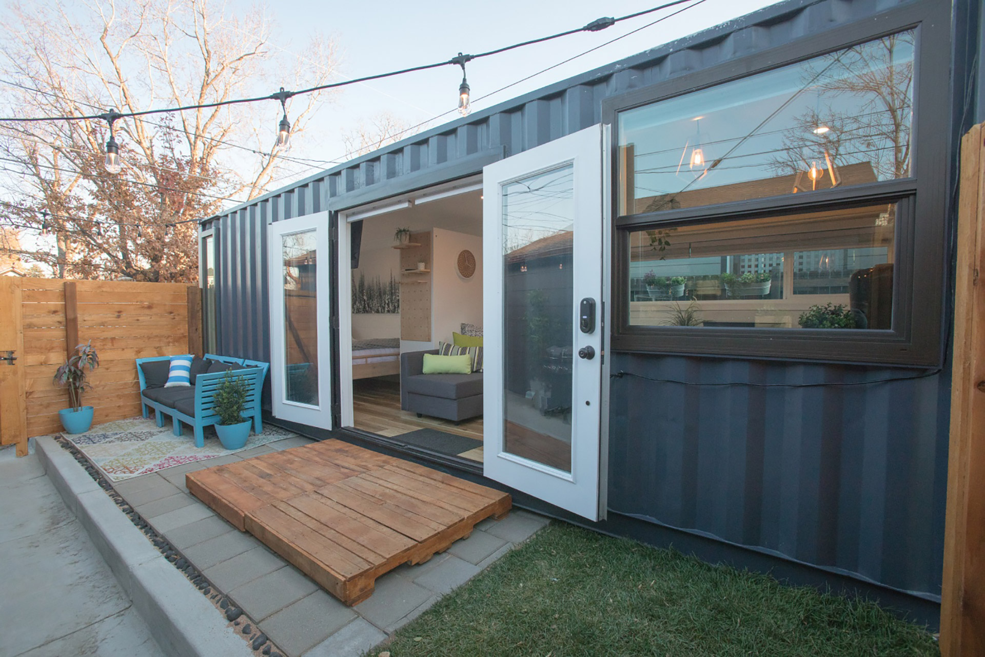 Modern guest house made from a dark gray shipping container with open white French doors, patio seating with blue furniture, and string lights overhead.