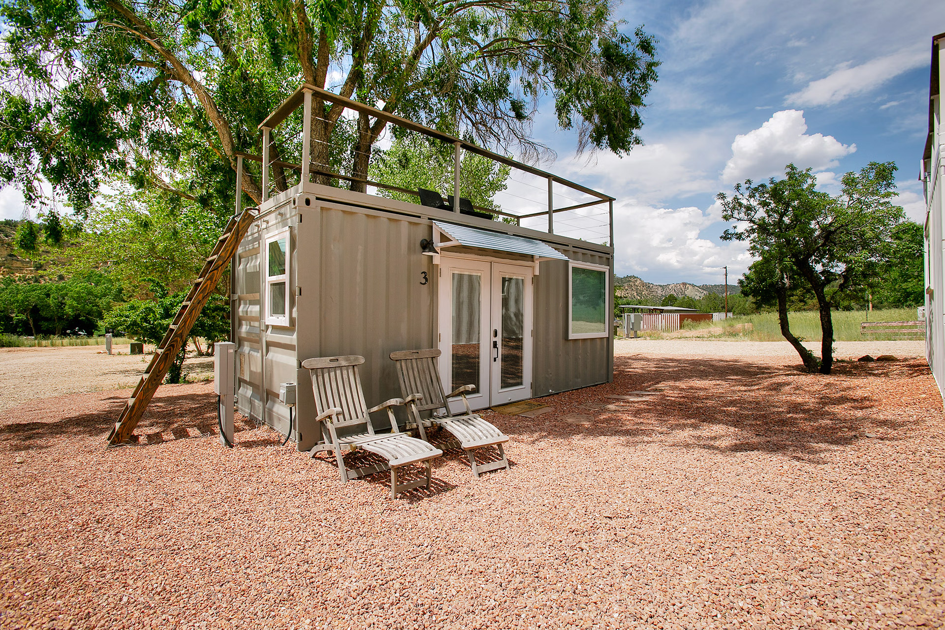 Small modern shipping container home with roof deck, wooden ladder, and two reclining chairs on gravel.
