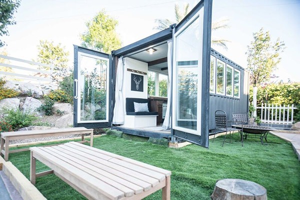 Small modern container house with open glass doors on a green lawn, surrounded by outdoor furniture and trees.