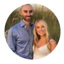 Two people smiling indoors with a decorated Christmas tree blurred in the background.