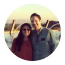 Smiling woman with blonde hair wearing a red scarf and a brown jacket, outdoors with sunlight in the background.