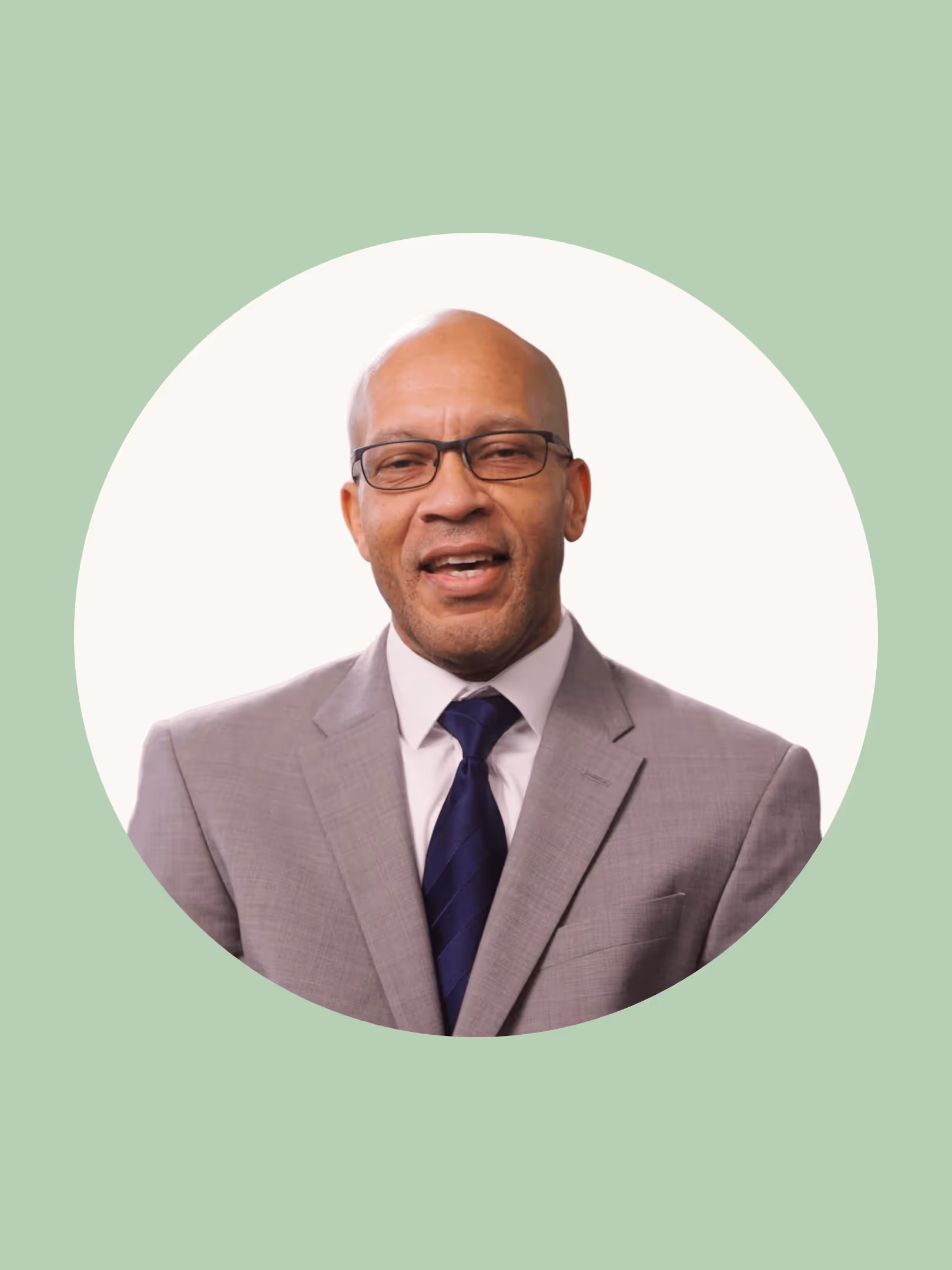 Smiling bald man wearing glasses, a gray suit, white shirt, and dark blue tie against a circular white background with green outer border.