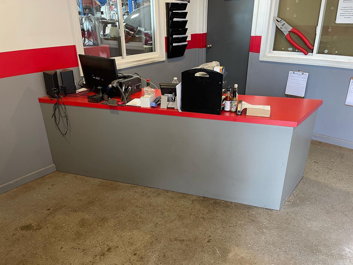 Reception counter with red countertop holding computer monitor, office supplies, hot sauce bottles, and black organizer in a room with gray and red walls.