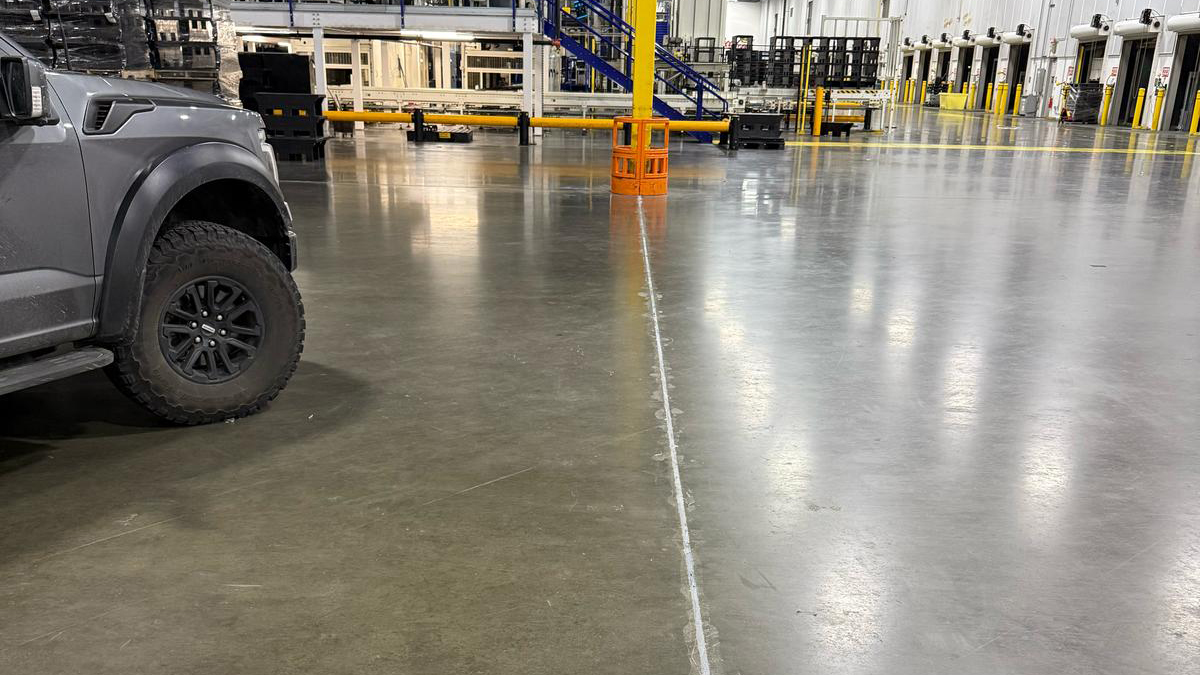 Industrial warehouse interior with polished concrete floors, yellow safety barriers, and a gray truck parked on the left.