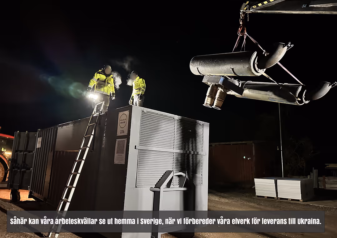 Två personer i varselkläder står på en container under natten, med en lyftkran som håller ett stort metallrör i förgrunden.