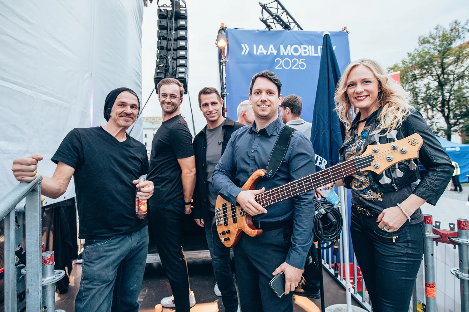 Five smiling band members posing backstage, one holding a bass guitar and another holding a drink bottle, with an IAA Mobility 2025 banner in the background.