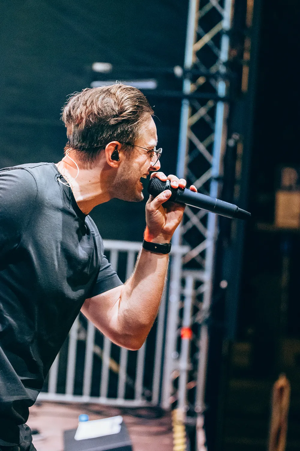 Male singer wearing glasses and a black t-shirt performing on stage with a microphone.