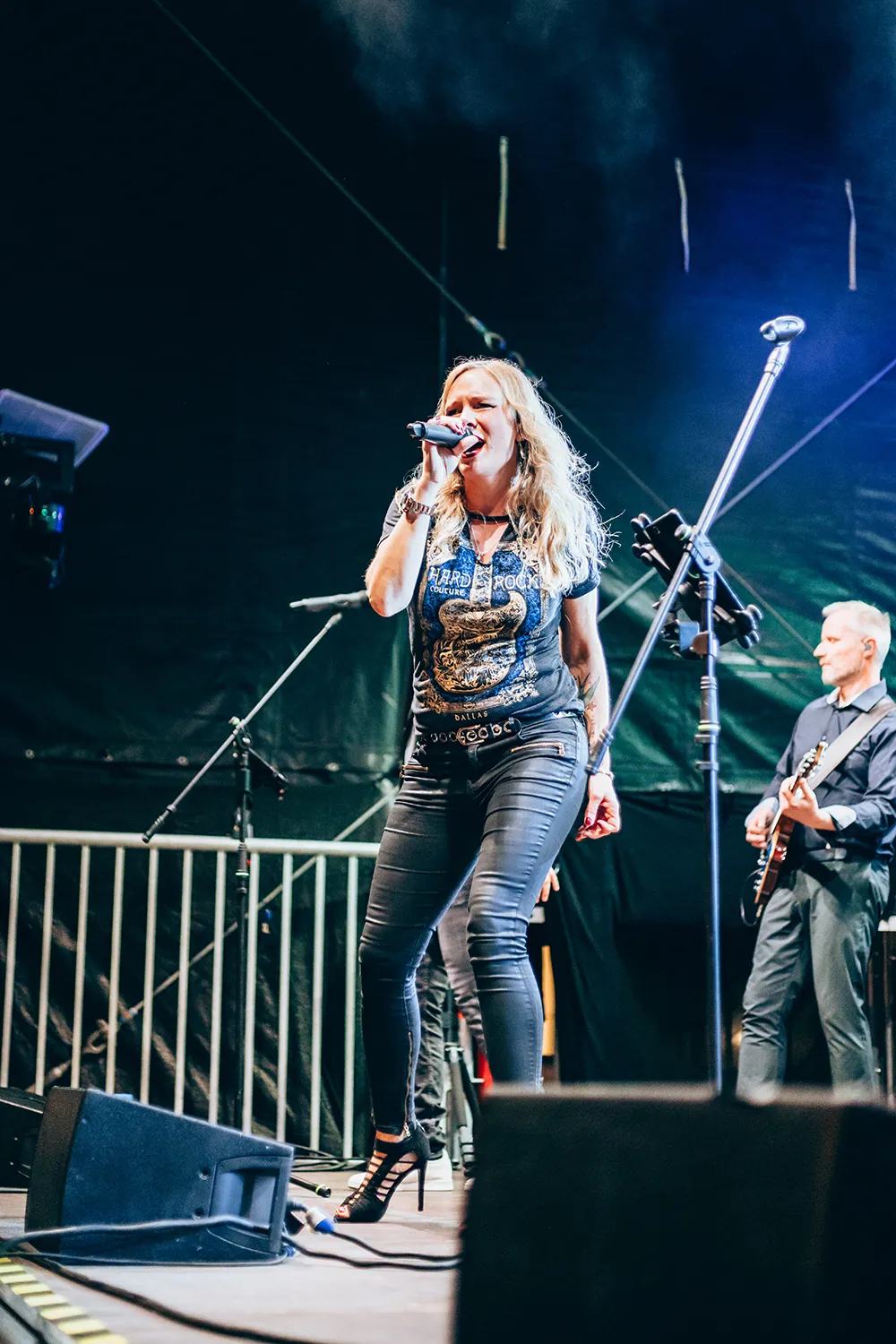 Female singer in black Hard Rock t-shirt and high heels performing on stage with guitarist in the background.