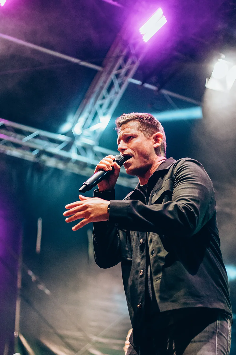 Male singer wearing a black jacket performing on stage with purple and blue stage lights overhead.