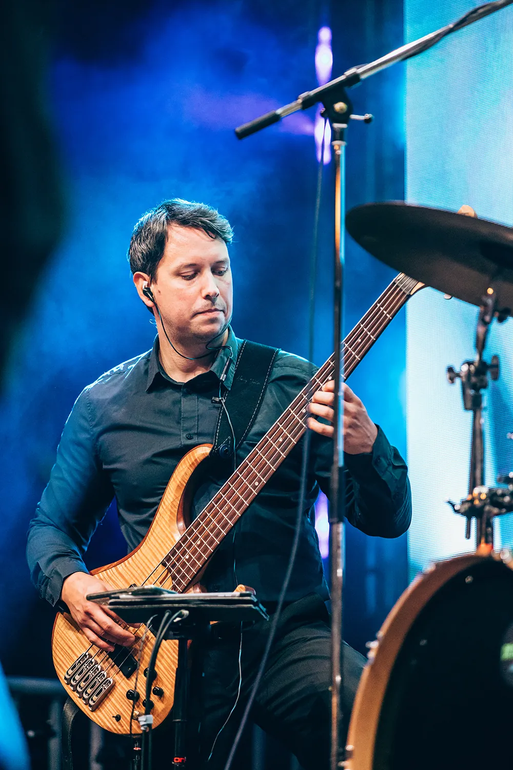 Musician wearing a black shirt playing a wooden electric bass guitar on stage with blue lighting and a drum set nearby.