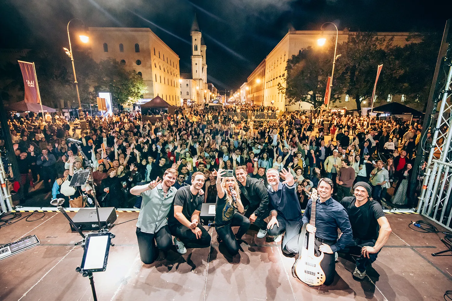 Musical band on outdoor stage taking a group selfie with a large crowd of cheering people behind them at night.