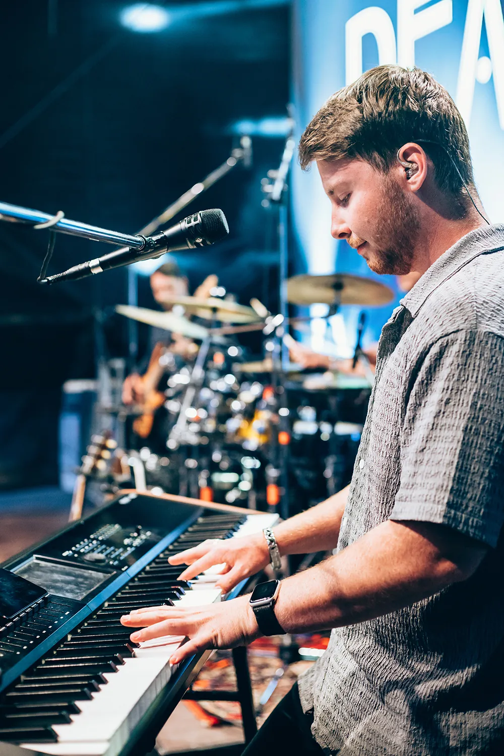 Man playing keyboard on stage wearing a gray shirt and smartwatch, with blurred drummer in the background.