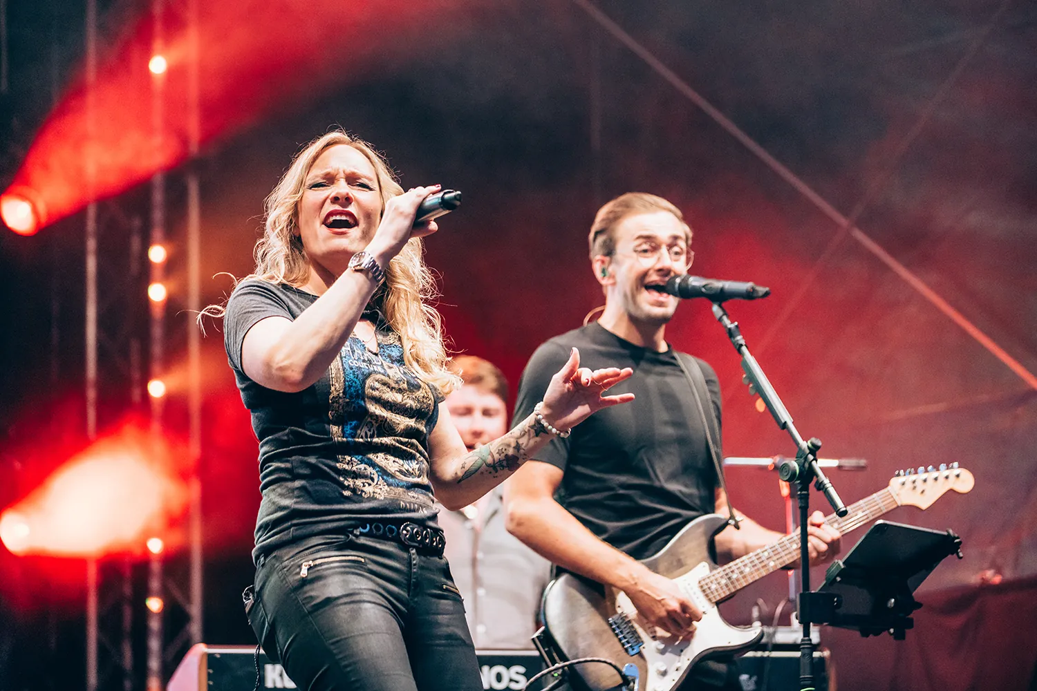 Female singer and male guitarist performing energetically on stage with red lighting.