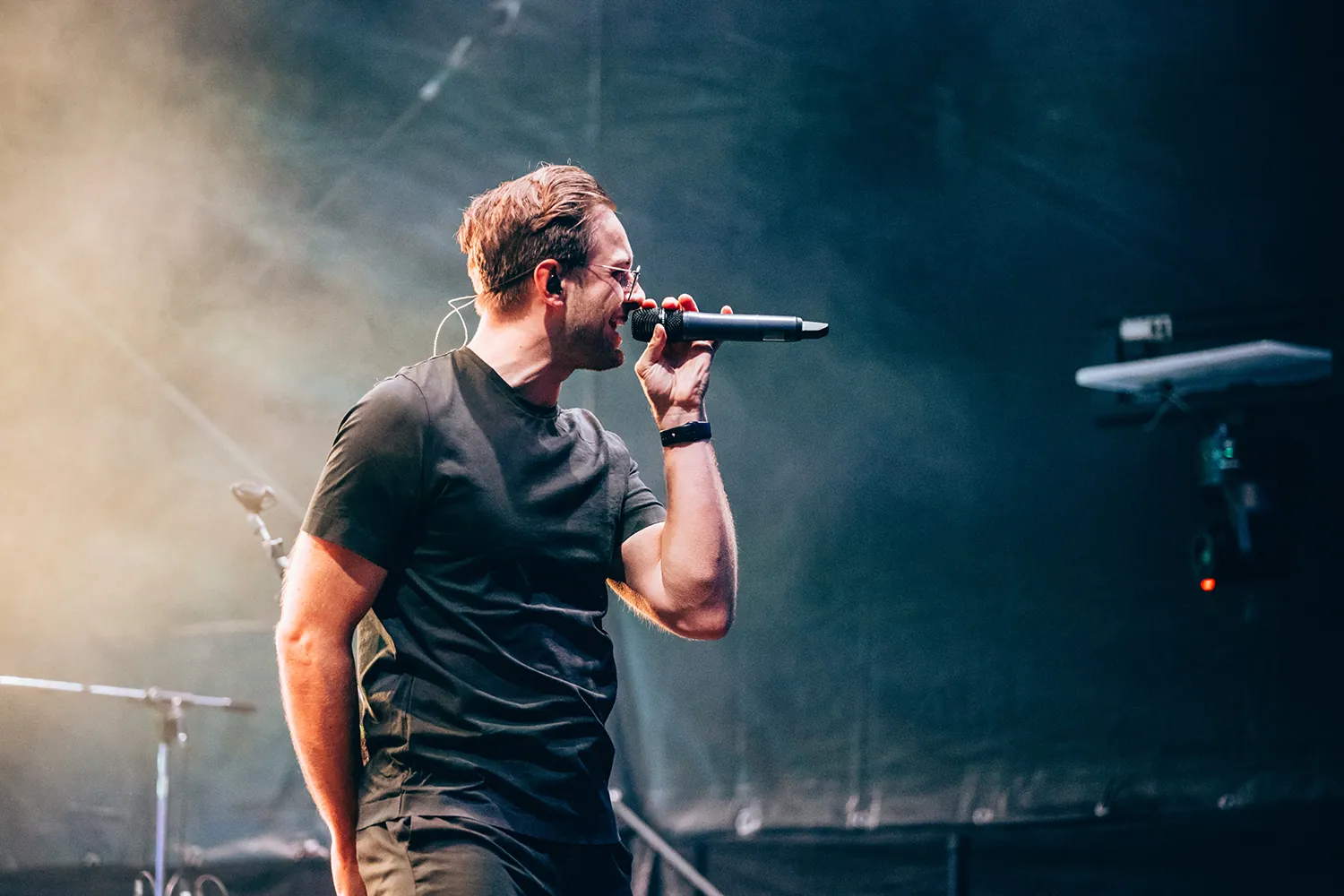 Male singer in black shirt holding a microphone and performing on stage with smoky lighting.