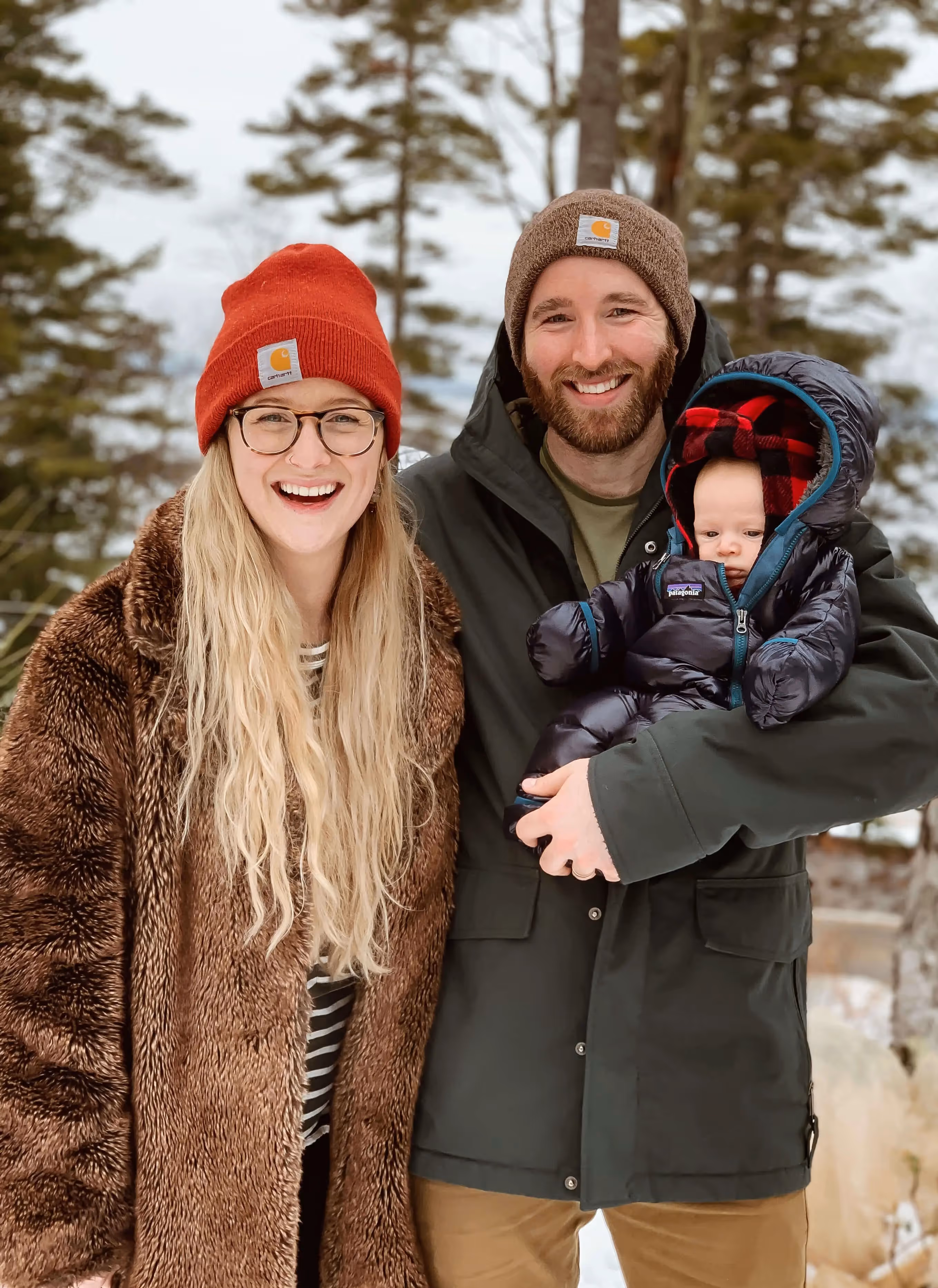 Family in winter coats and Carhartt beanies smiling in snowy forest