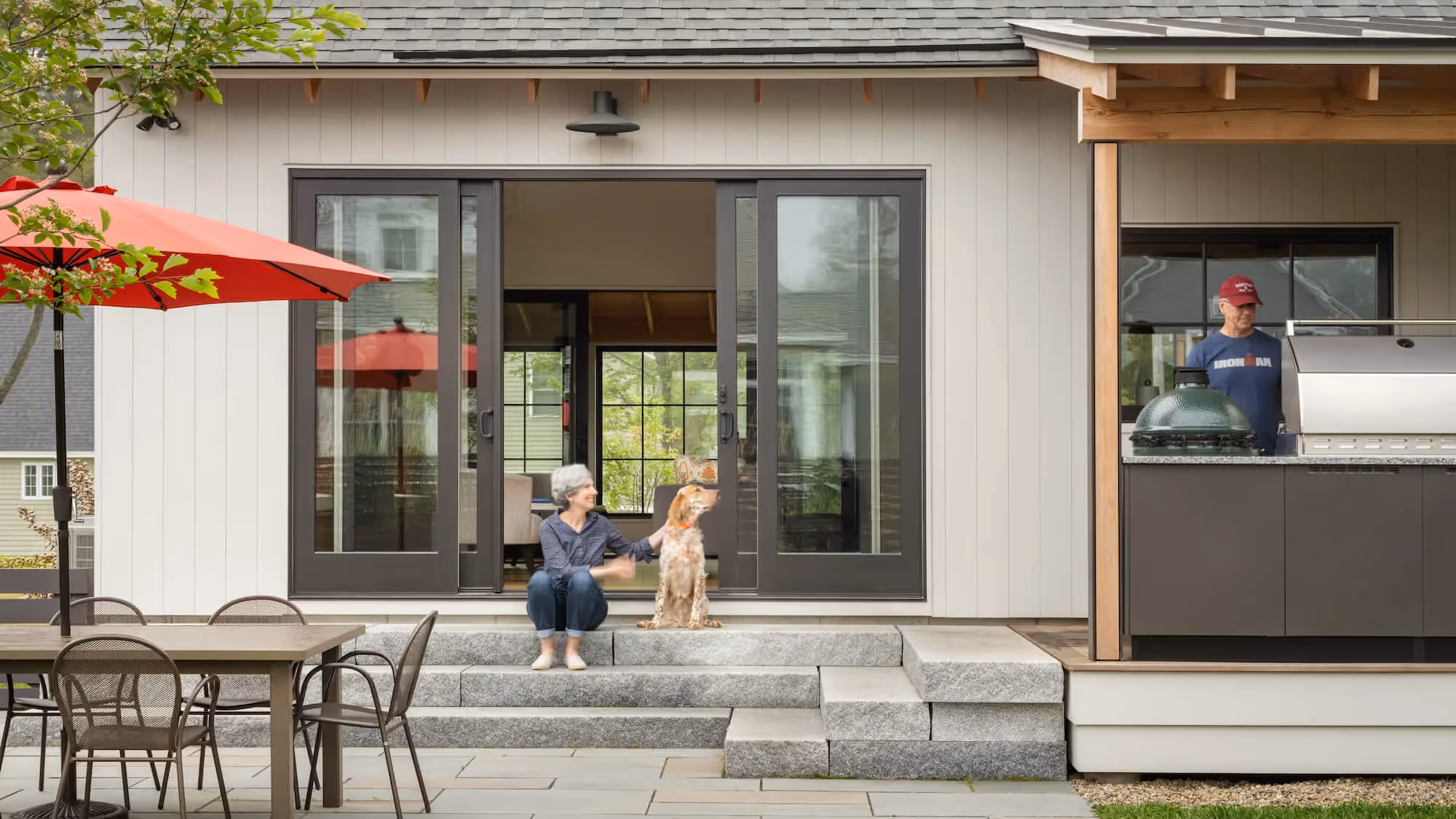 Person sitting with dog on patio steps near outdoor kitchen and table