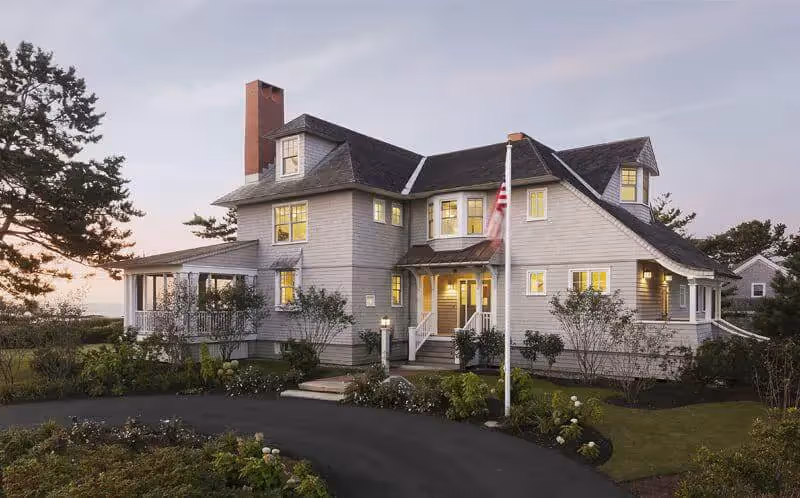 Twilight view of a large white coastal home with warm glowing windows