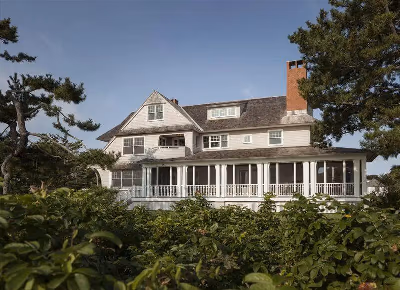Large white coastal home with wraparound porch surrounded by green foliage