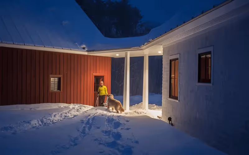 Person in yellow jacket with dog outside snowy red wooden house at night