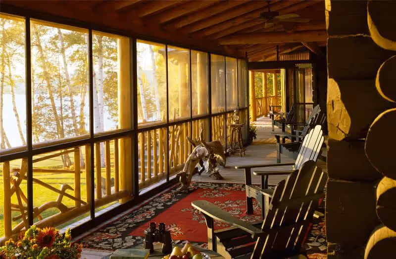 Sunlit wooden porch with Adirondack chairs overlooking forest through large windows