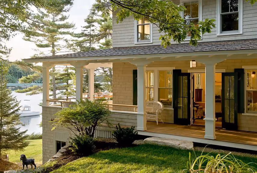 Lakeside home with white porch, green trees, and small dog on lawn