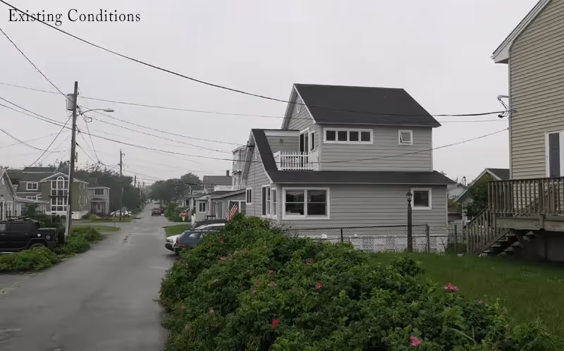 Residential street with gray houses, power lines, and green bushes with pink flowers