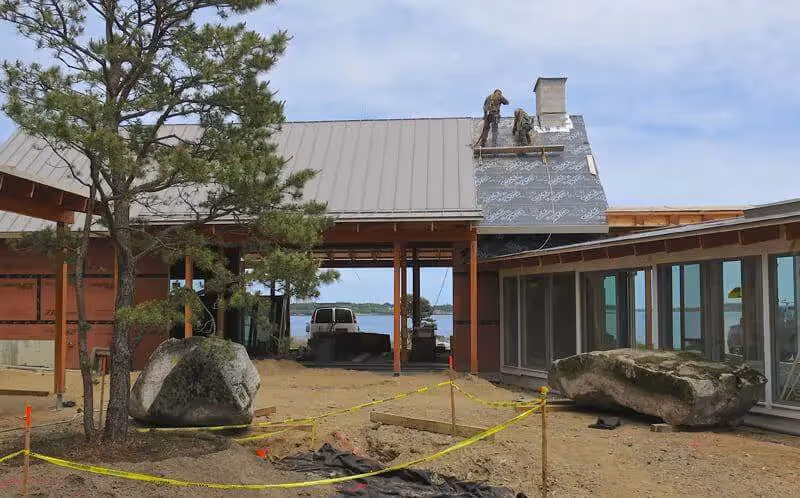 Workers installing metal roof on coastal building with large boulders nearby