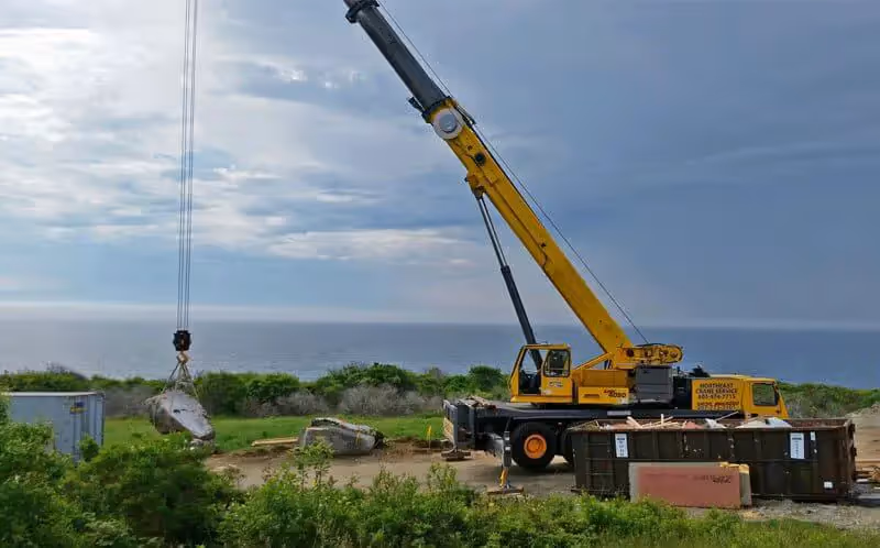 Yellow mobile crane lifting debris near ocean with cloudy sky