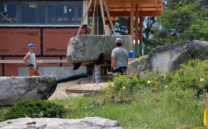 Workers using crane to move large boulder at construction site with greenery