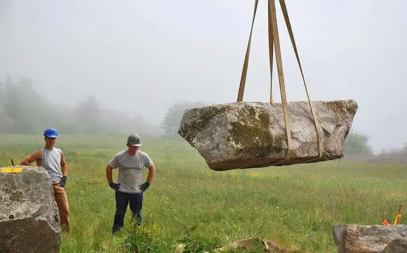 Large boulder suspended by straps with workers in misty grassy field
