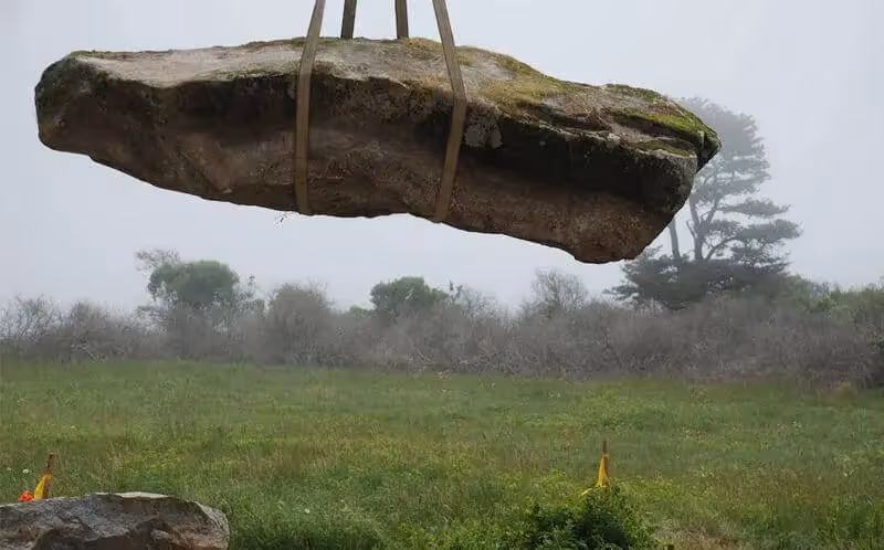 Large mossy boulder suspended by straps over misty green field