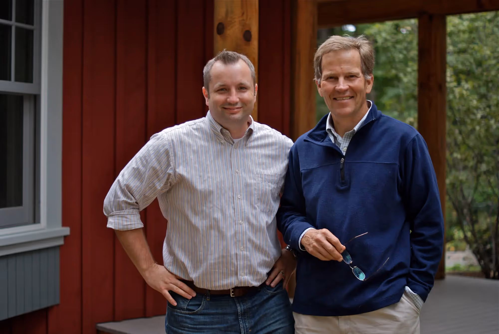 Two men standing together on a porch with red wooden building behind