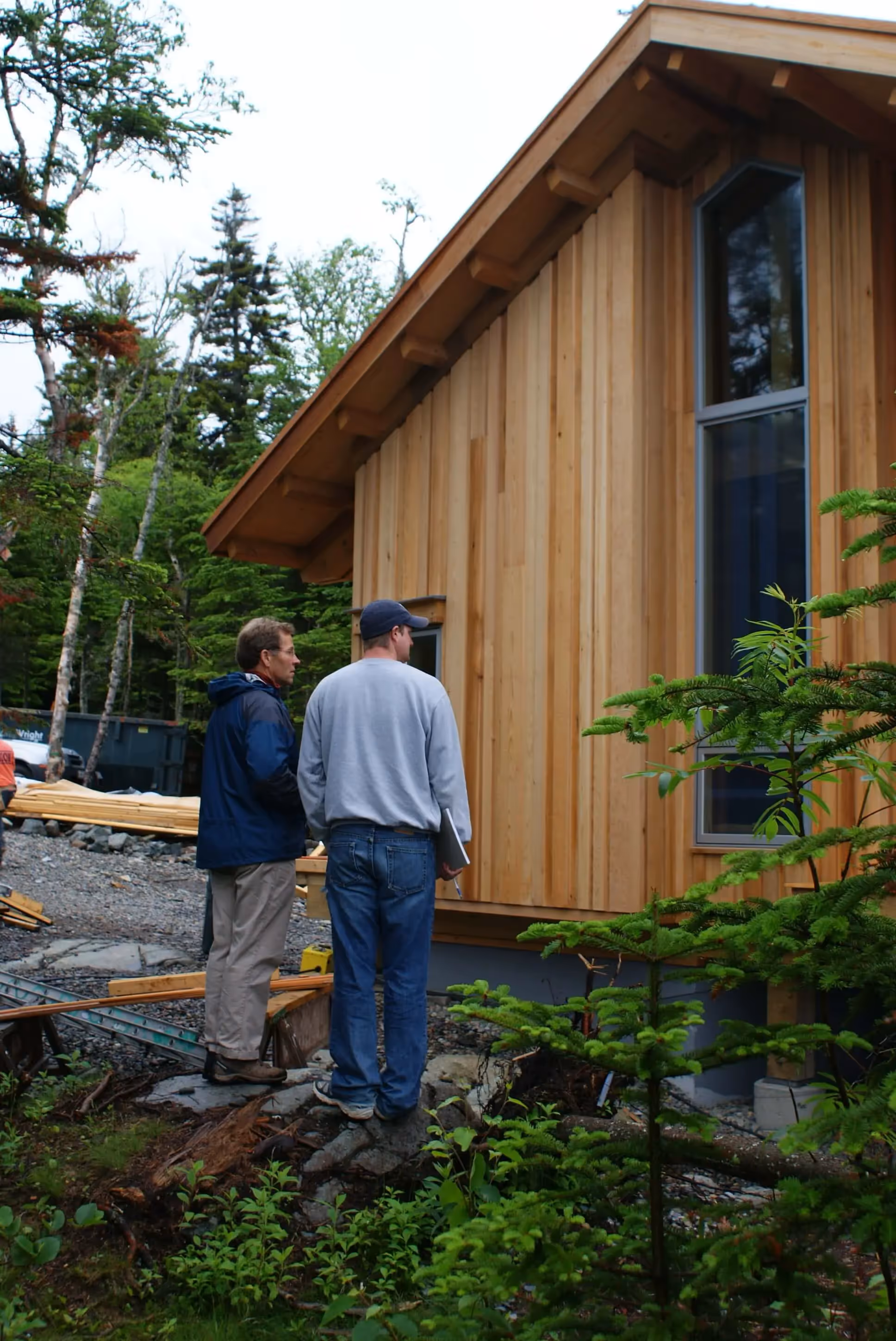 Two men inspect wooden cabin construction surrounded by forest trees