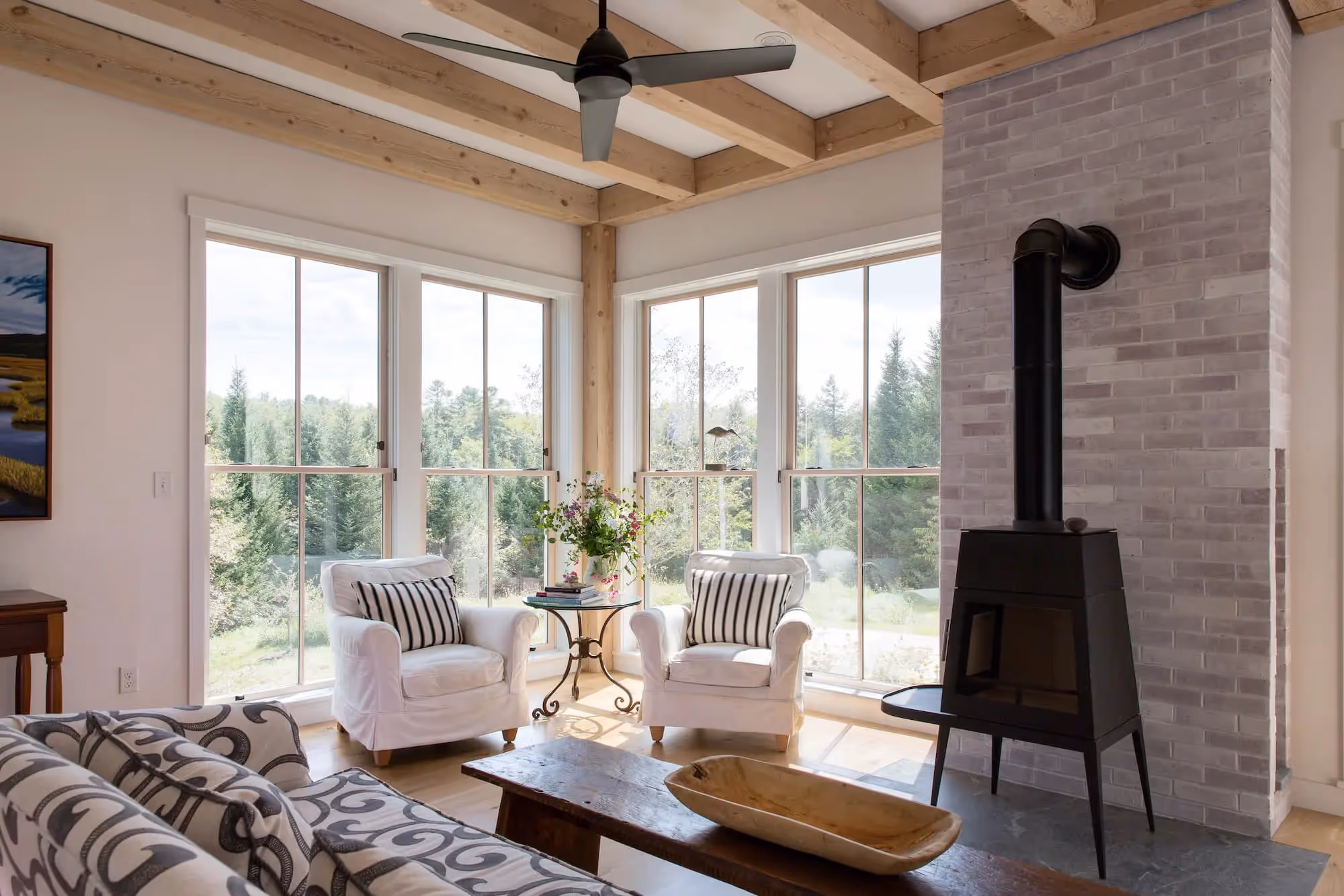 Cozy living room with large windows, white chairs, and wood stove