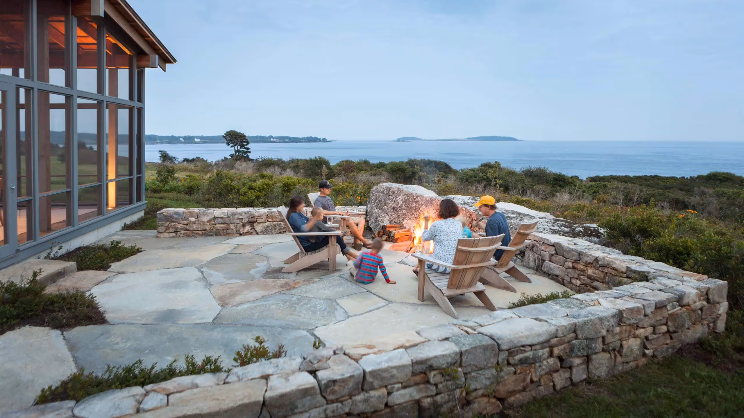 Family gathering around stone firepit with ocean view at sunset