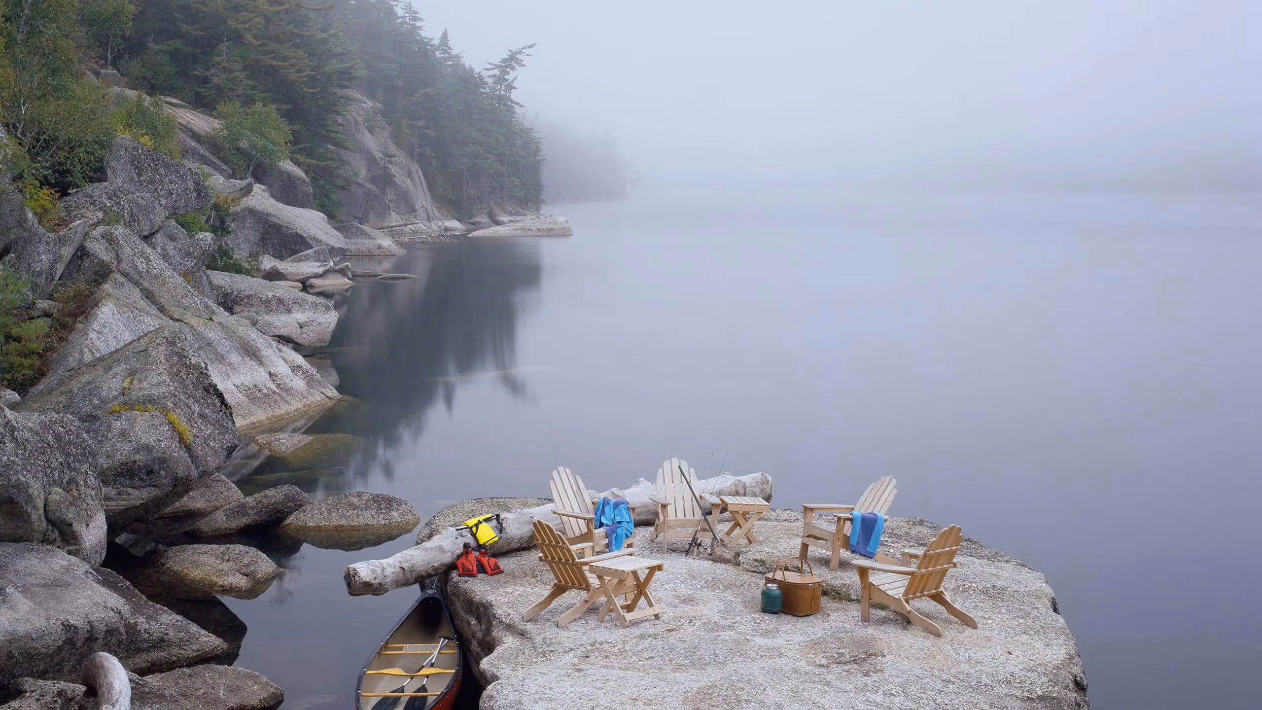 Misty lake with Adirondack chairs on rocky shore, canoe below