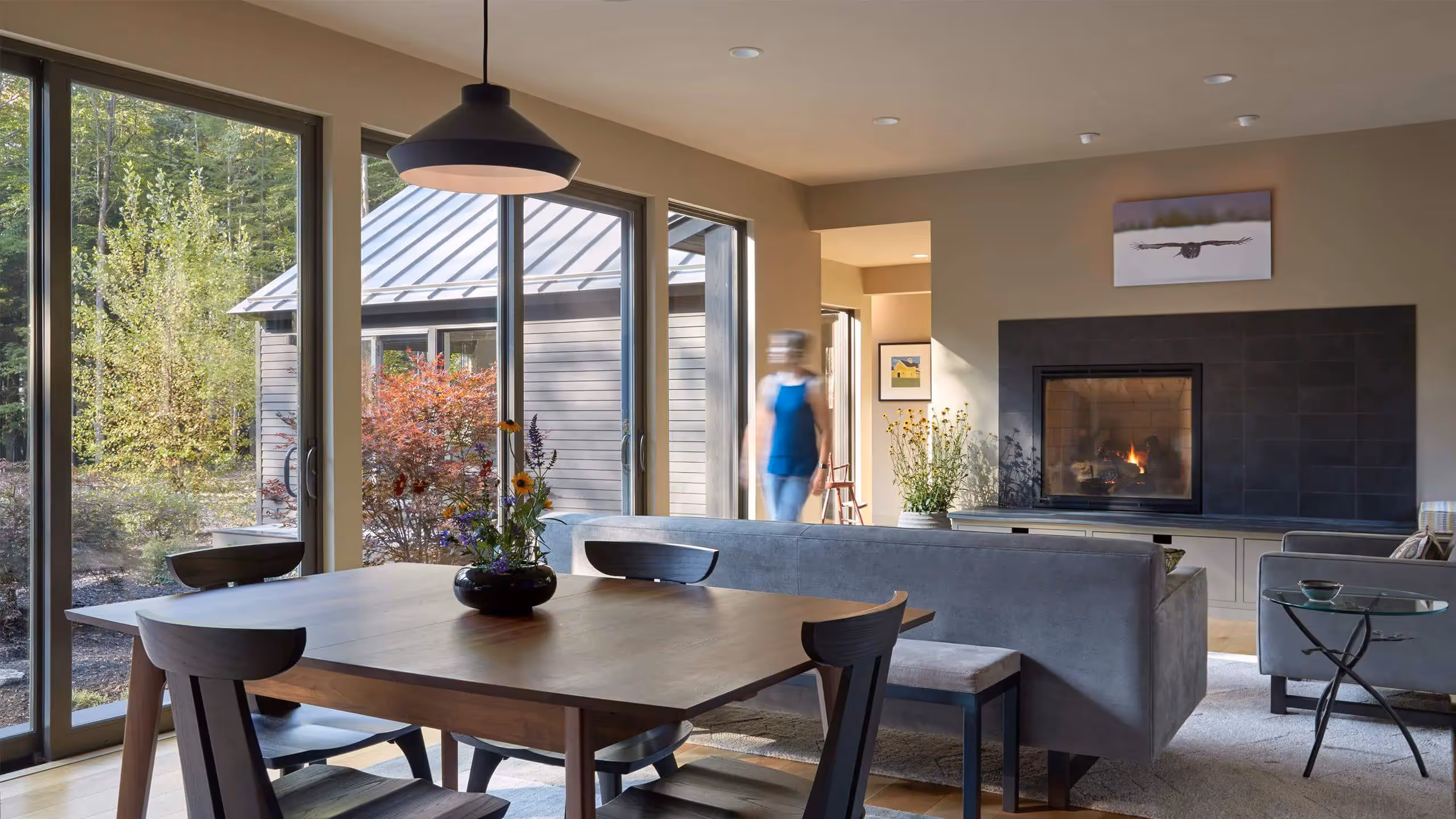 Modern dining area with large windows, fireplace, and blurred figure walking
