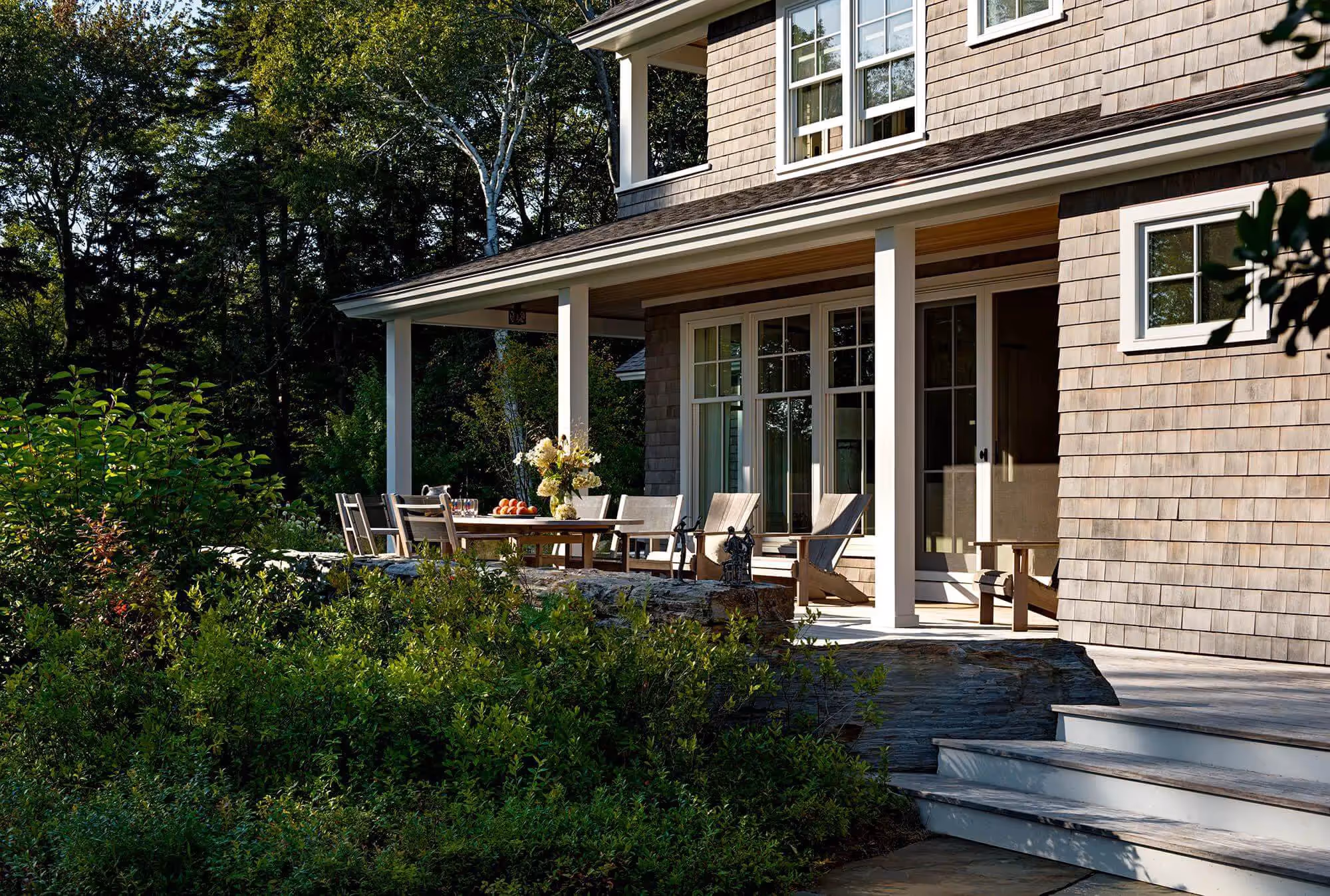 Shingle-style house with porch, outdoor dining table, and lush green landscaping