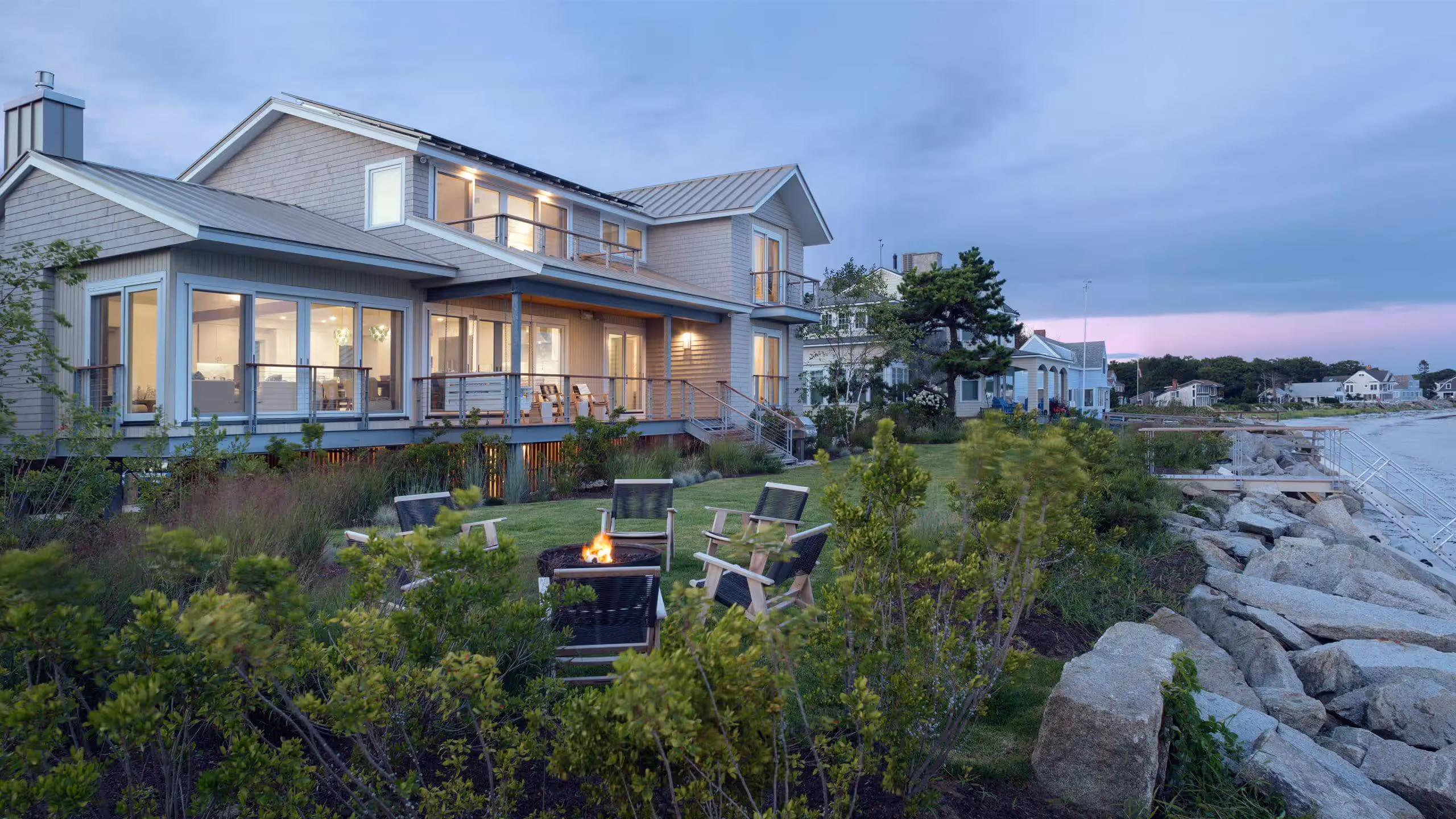 Coastal beach house with fire pit at dusk, overlooking rocky shoreline