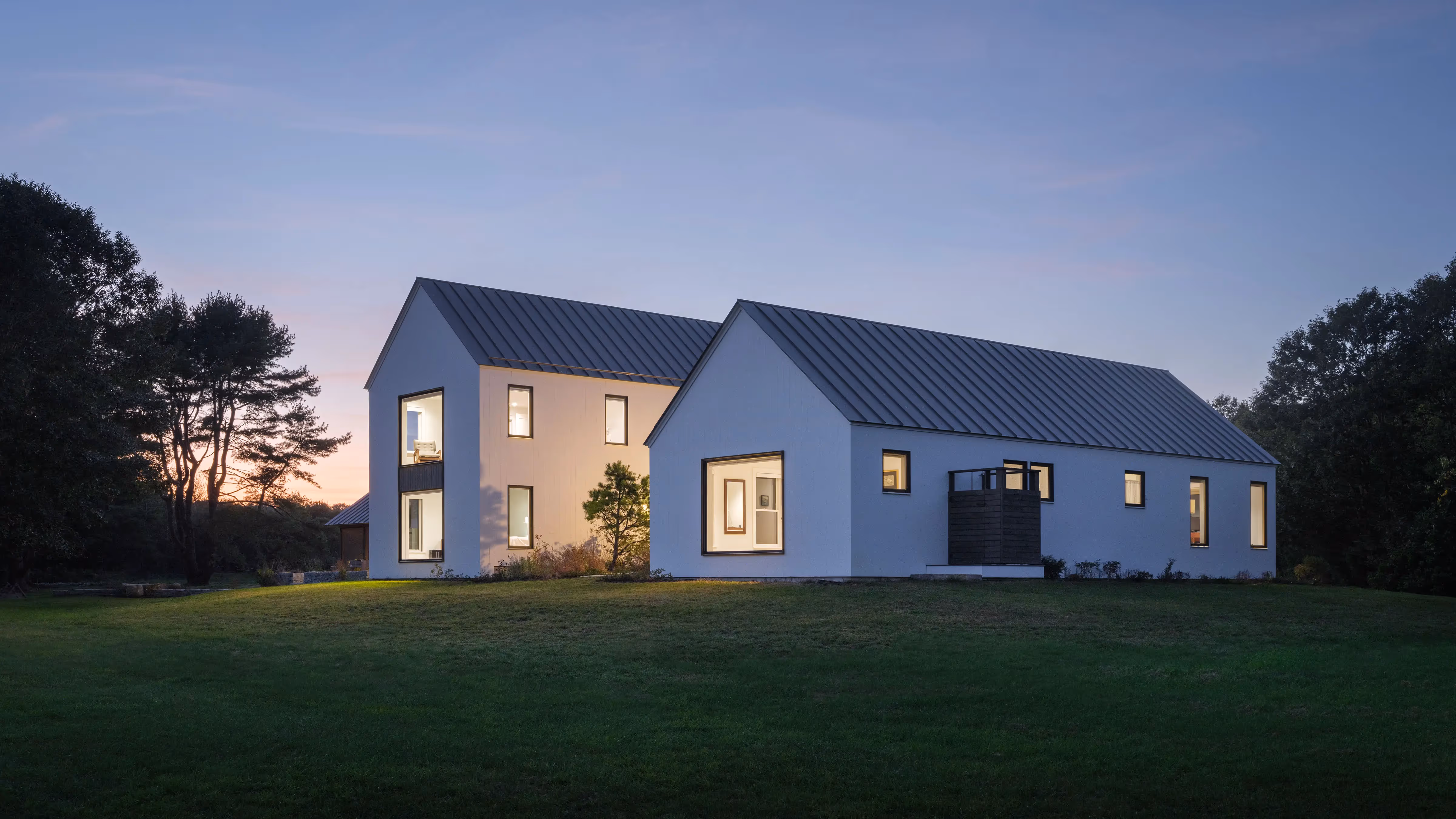 Modern white house with glowing windows at dusk, surrounded by green lawn